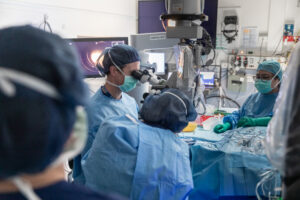 Opthalmic surgeons and theatre staff working on a gene therapy trial. The lead surgeon is looking through a piece of opthalmic equipment and are shown from the back. All are wearing blue surgical gowns, caps and masks.