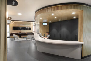 A large desk in the lobby of waiting room. There is a person sitting behind the desk, and behind them some patients sitting