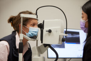 A woman wearing a surgical mask is looking through an ophthalmic device in a clinical setting. A female clinician (mostly out of frame) sits opposite and is also wearing a mask.