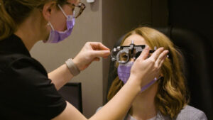 A woman, wearing a purple face fask, has a pair of refractor glasses put on her face by a clinician