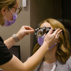 A woman, wearing a purple face fask, has a pair of refractor glasses put on her face by a clinician