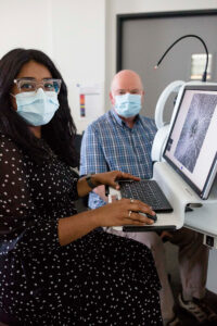 A clinician operates a computer while a patient sits near an ophthalmic camera in the background