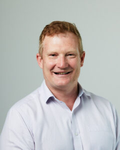 Portrait image of Darren Kelly taken against a plain grey background. He is wearing a white shirt.