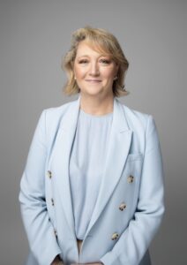 Image of Michelle Gallaher standing in front of a grey backgorund. She is wearing a pale blue suit.