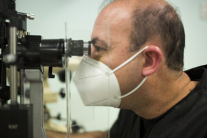 Image of clinical trial worker in black scrubs and a nmask looking into a slit lamp.