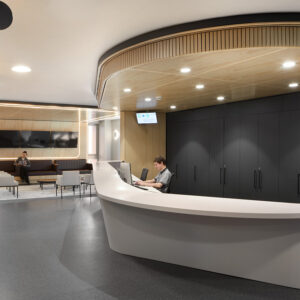 A large desk in the lobby of waiting room. There is a person sitting behind the desk, and behind them some patients sitting