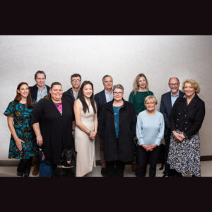 A group of 11 people are standing together in two rows and smiling at the camera in front of a light grey wall.
