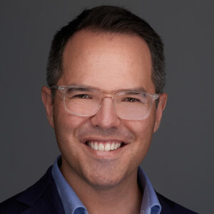 Headshot of Nick Northcott. He is smiling and wearing a black jacket, blue shirt and clear-framed glasses.