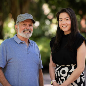 An older man with a grey beard and a cap stands next to a younger woman with long dark hair. Both are smiling and standing outdoors with greenery in the blurred background. The man is wearing a blue polo shirt, and the woman is wearing a black top and a black and white patterned skirt.