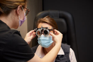 A clinician adjusts a metal eye-testing trial frame on a seated person wearing a face mask, inside a dark exam room with a padded examination chair.
