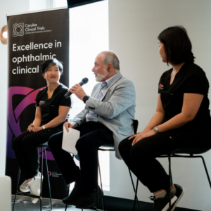 Three panelists seated on stools at an indoor event; the person in the centre speaks into a handheld microphone while the two others listen. A vertical banner behind them reads “Cerulea Clinical Trials – Excellence in ophthalmic clinical,” with a purple graphic. The panelists wear dark polo shirts with logos, and the setting appears to be a bright conference or seminar space.