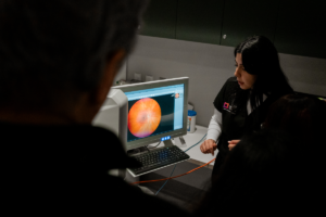 A healthcare professional stands beside a desktop computer displaying a close-up retinal scan image while explaining the screen to two people nearby; the scene appears to take place in a dimly lit clinical exam room with medical equipment visible in the background.
