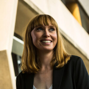 Professional portrait of Dr Jennifer Hollands, smiling and looking to the right of frame. A modern building appears close up in the background.